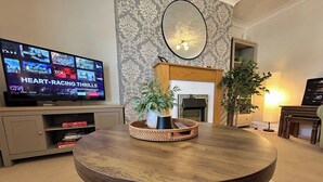 A round wooden table with a woven basket centerpiece, a flat-screen TV displaying various shows, a fireplace, and patterned wallpaper. - Central Bedford Apartment | Secure Parking | Fast Wi-Fi | Walk to Station (Bedford)
