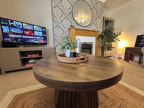 A round wooden table with a woven basket centerpiece, a flat-screen TV displaying various shows, a fireplace, and patterned wallpaper.
