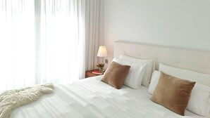 Ett hotellrum med en säng, vita gardiner, en sänglampa och ett fönster med tunna, genomskinliga gardiner.