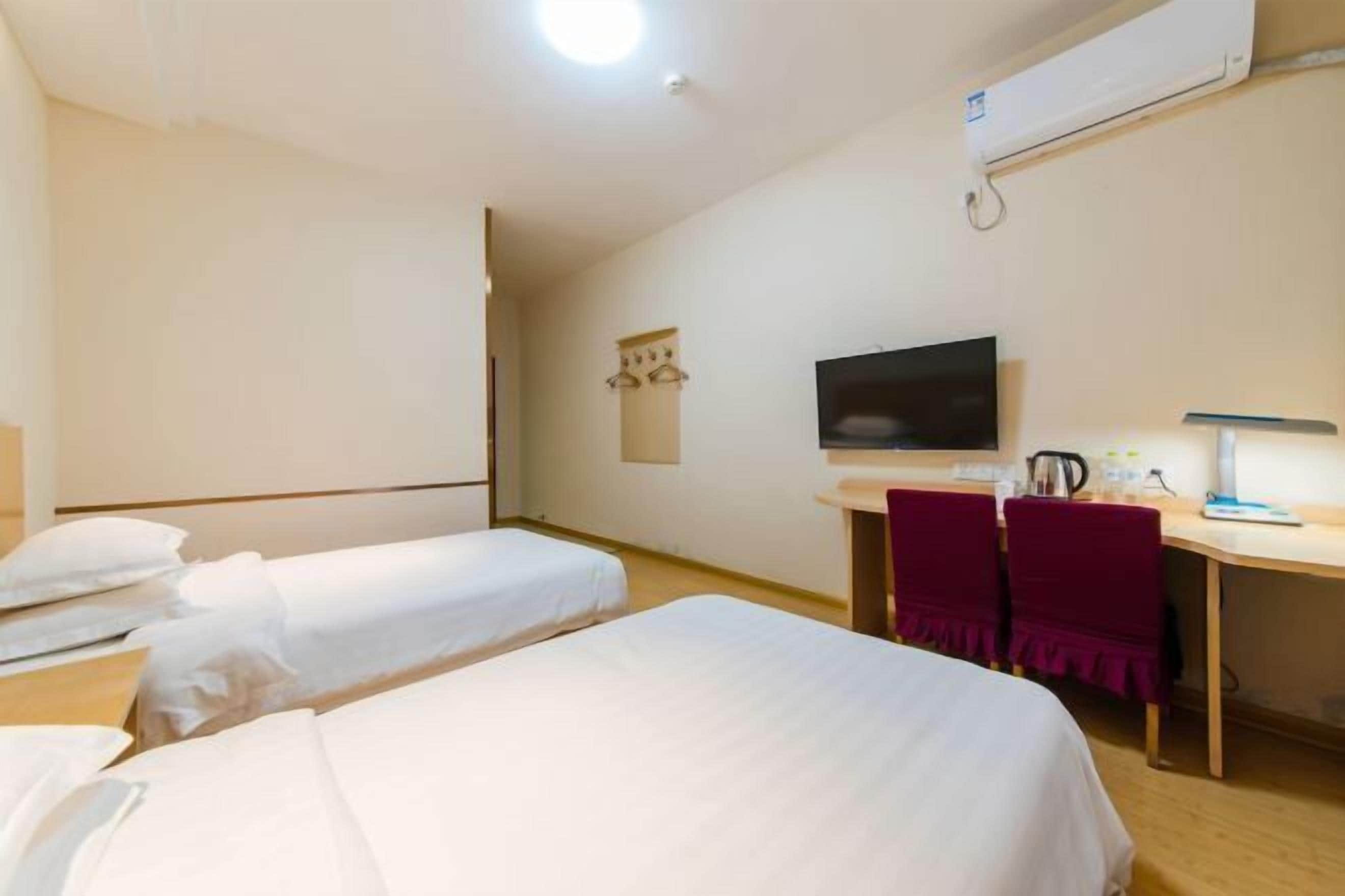 A hotel room with two single beds, a desk with a chair, a television, and an air conditioning unit.