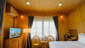 A room with a bed, two wicker chairs, a small table with a lamp, a television on a stand, and a window with curtains. - Cantho Eco Resort (Can Tho)