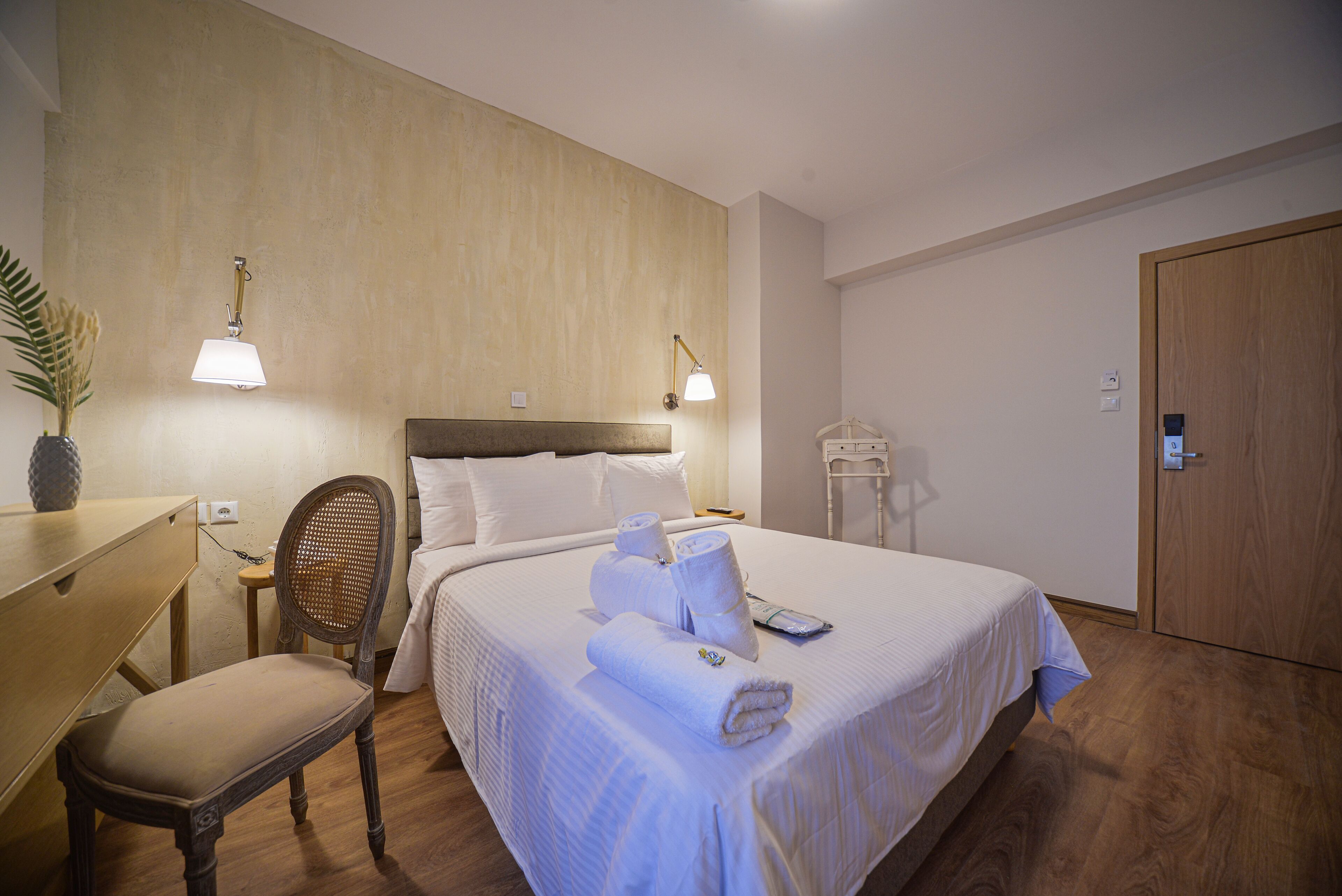 Quarto de hotel com cama, escrivaninha, cadeira e duas luminárias fixadas na parede.