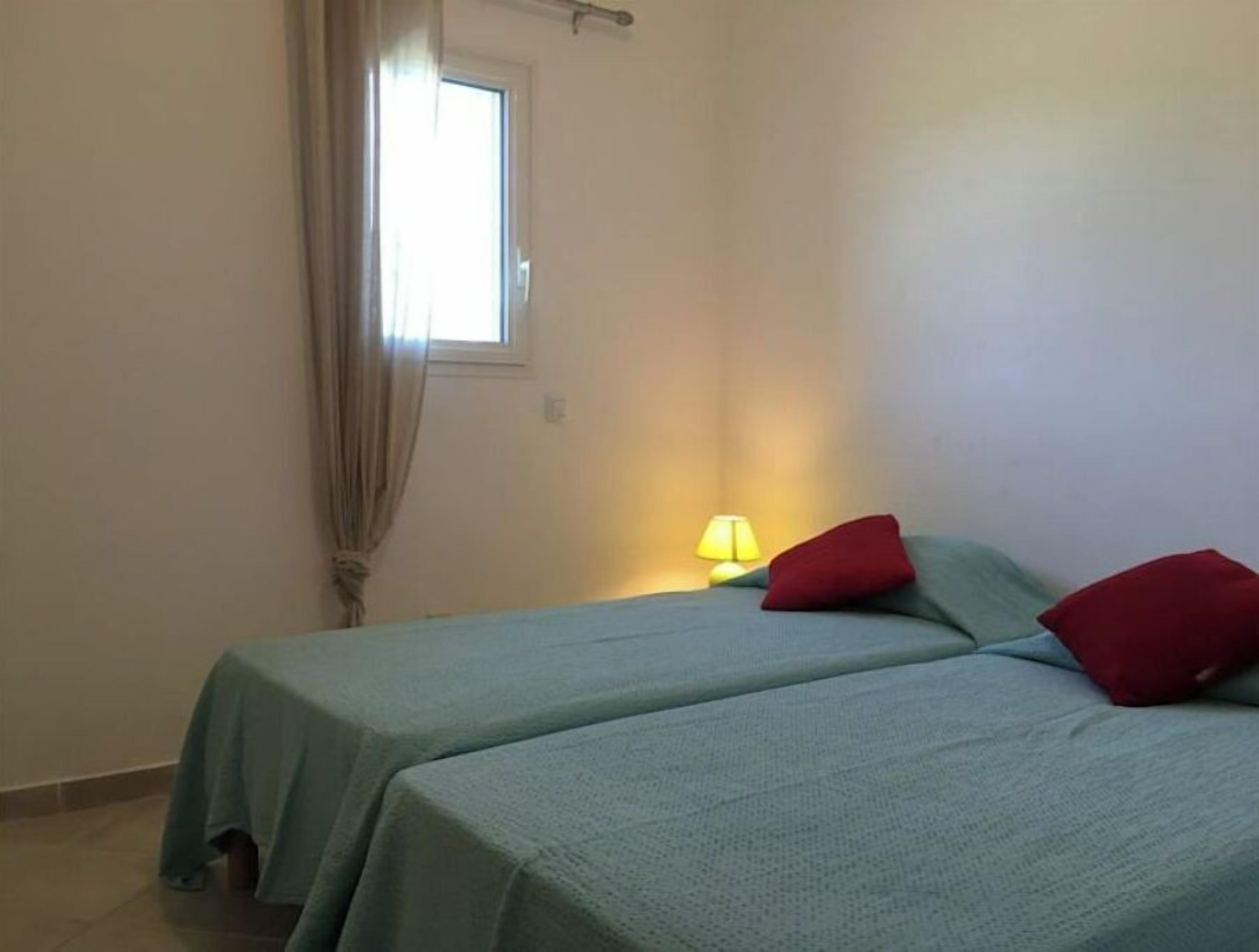 Una habitación pequeña y sencilla con dos camas, una ventana con cortinas y una lámpara de noche.