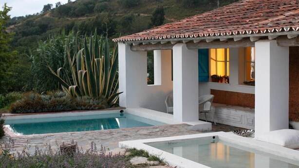 Una piscina con exterior blanco, un área de patio con una silla blanca y vistas a un paisaje montañoso.