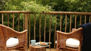 Dos sillas de mimbre con cojines blancos sobre una terraza de madera, una pequeña mesa con una taza y un plato de comida, y una valla de bambú al fondo.