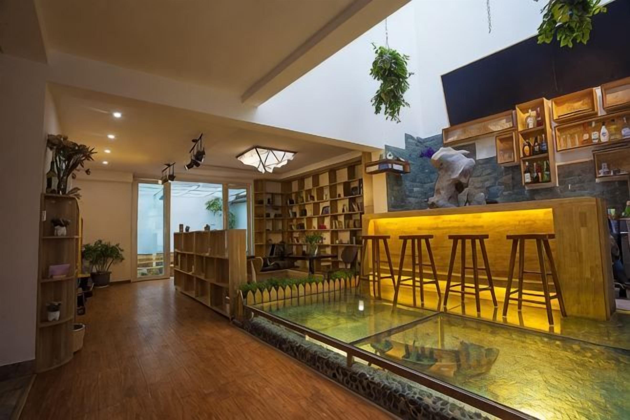 Un espacio interior amplio con piso de madera, una mesa de vidrio y una zona de bar con taburetes. Hay estantes con varios objetos y un gran ventanal que proporciona luz natural.