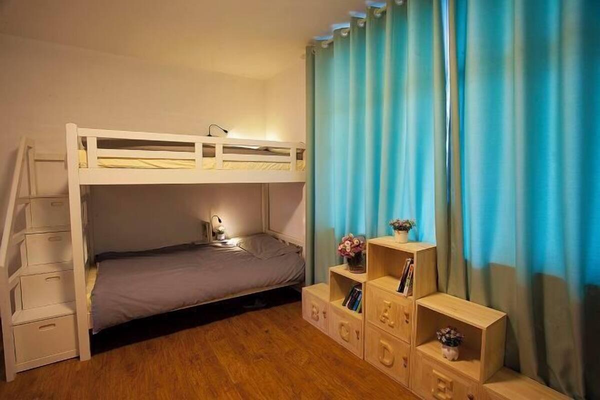 A bunk bed room with a desk and drawers, a lamp, and a bookshelf.