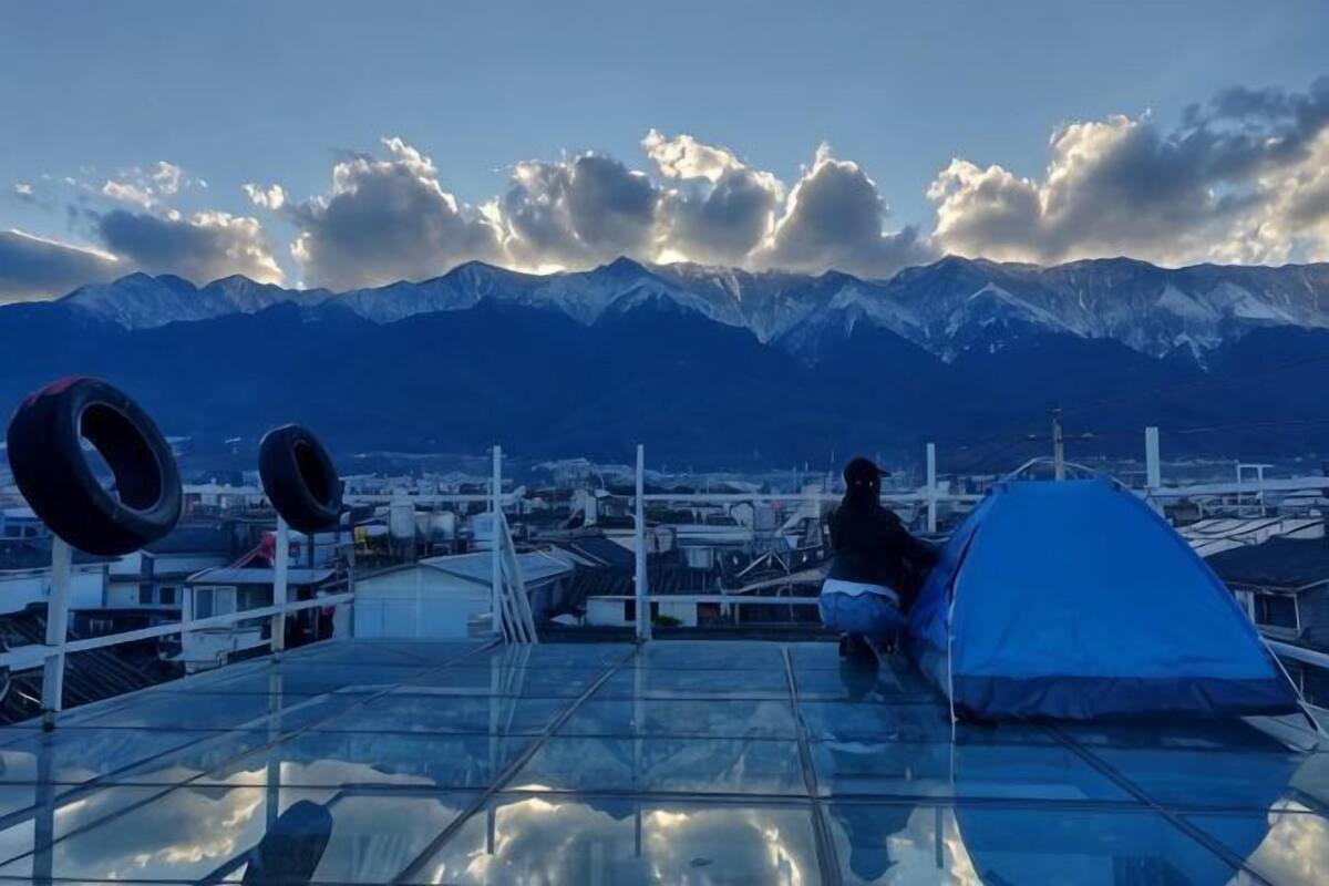 A person sitting on a rooftop with a tent and tires, overlooking a city and mountains.