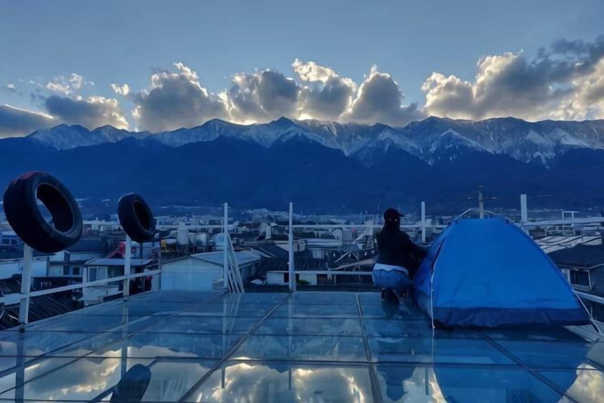 A person sitting on a rooftop with a tent and tires, overlooking a city and mountains.