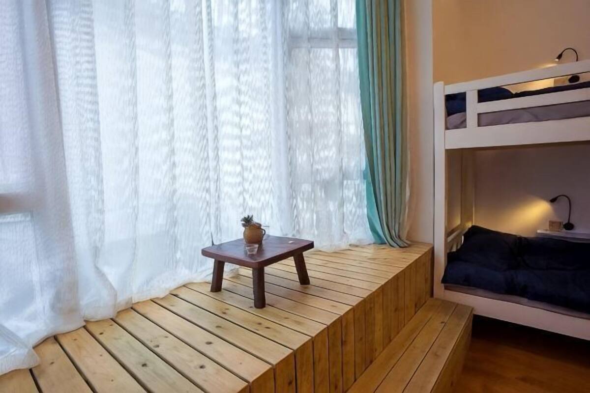 A bunk bed with a small table and a plant, a window with sheer curtains, and a wooden bench.