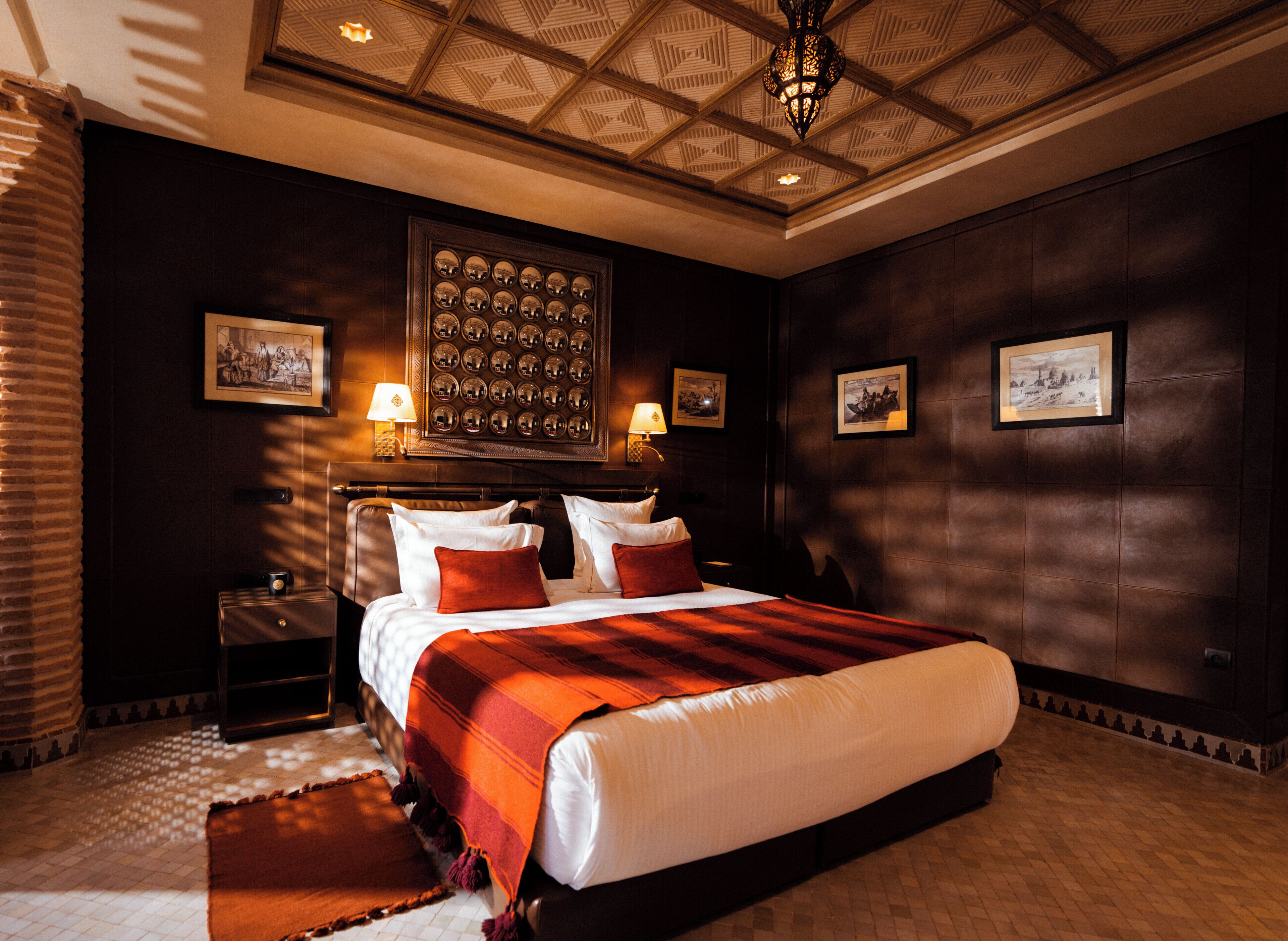 A large, dark-themed bedroom with a large bed, multiple pillows, a headboard with a decorative mirror, and a patterned ceiling.