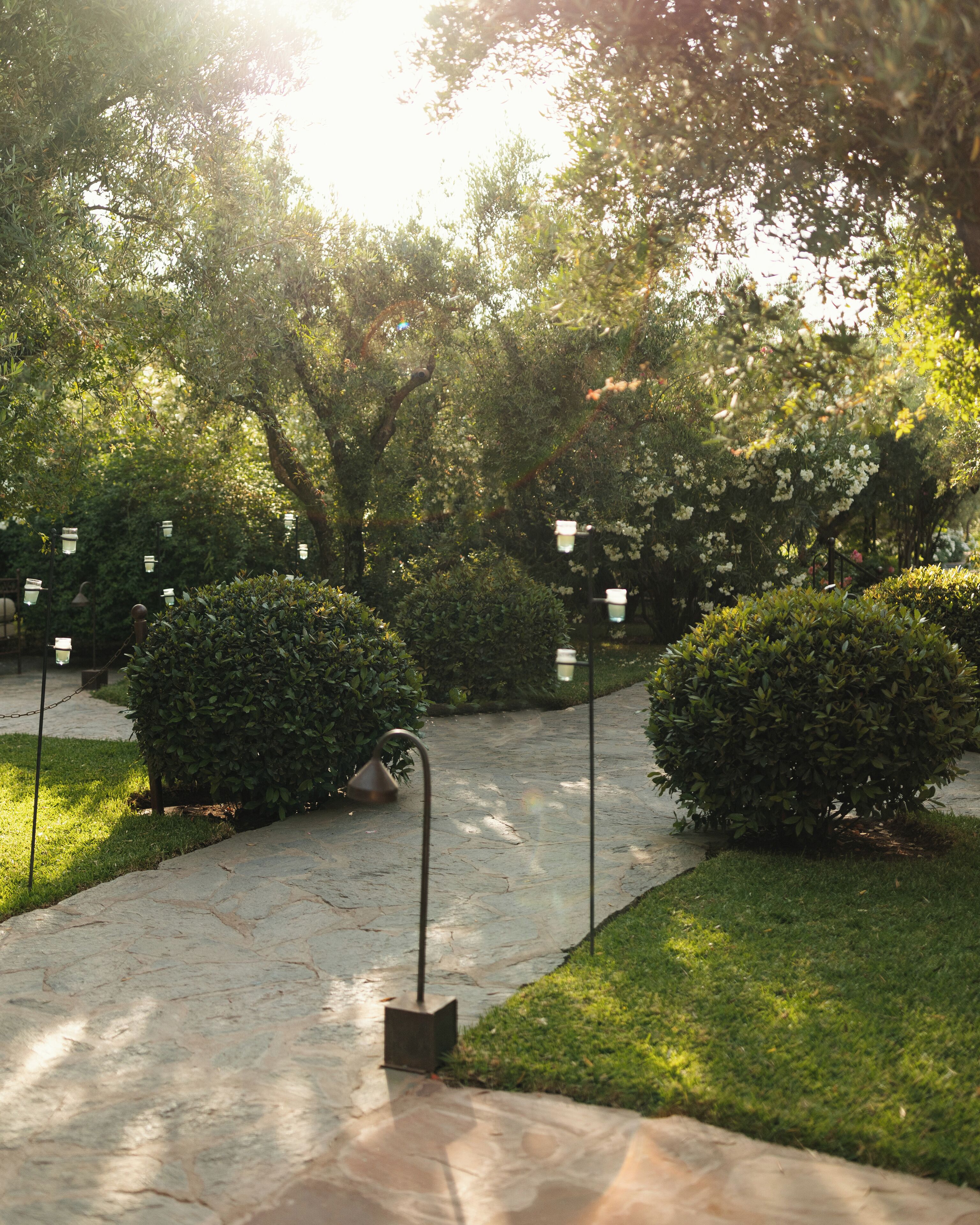 A stone pathway in a park with neatly trimmed bushes and trees, surrounded by a well-maintained lawn.