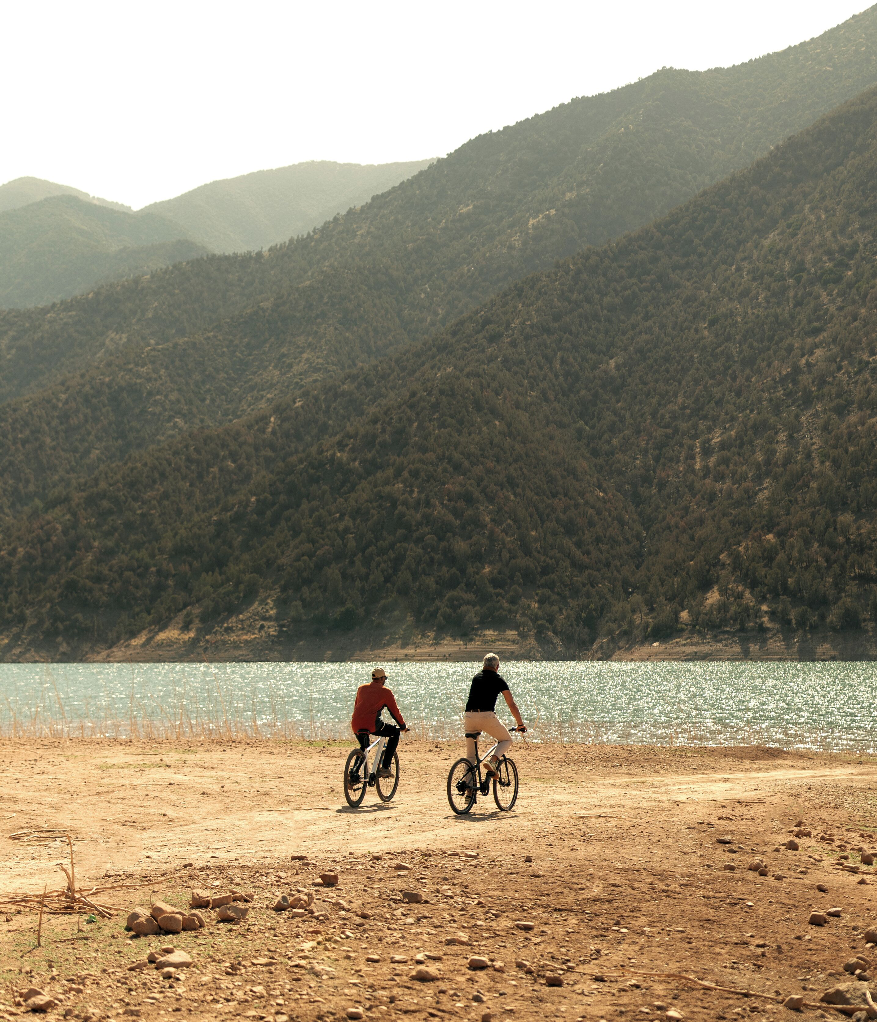 Deux personnes font du vélo sur un chemin de terre près d’une étendue d’eau, avec des collines en arrière-plan.