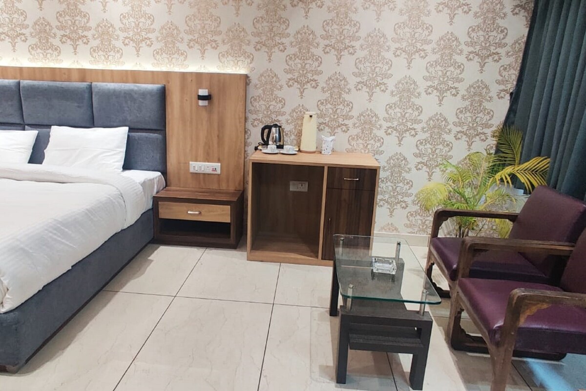A hotel room with a bed, a glass table, a purple armchair, a wooden cabinet, and a potted plant.