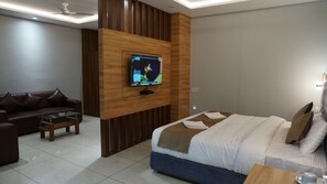 A modern hotel room with a large bed, a sofa, a TV mounted on a wooden panel, and a ceiling fan. - HOTEL SATKAR (Katihar)