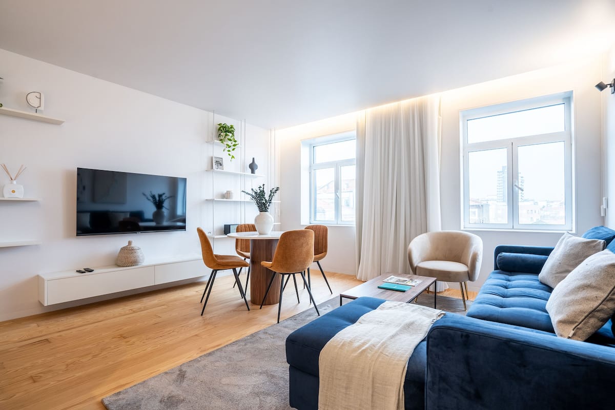 Una sala moderna con un sofá azul, un televisor, un comedor con sillas y un amplio ventanal con cortinas.