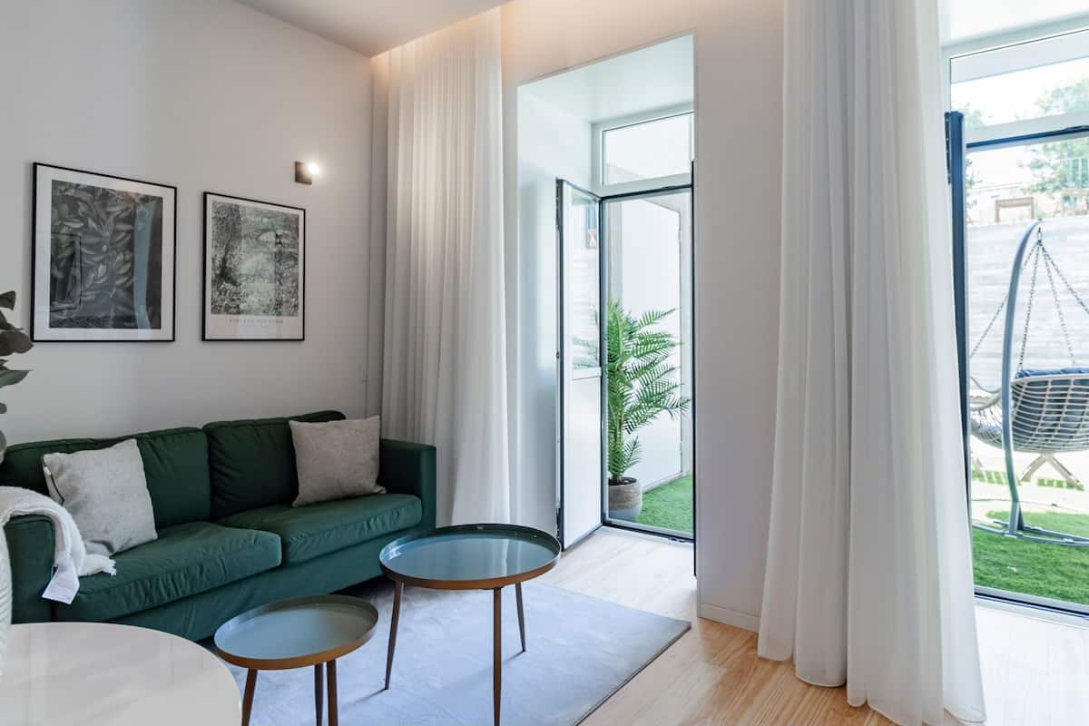 Una sala moderna con un sofá verde, dos mesas con superficie de vidrio y un sillón colgante junto a la ventana.