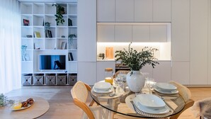 A modern dining area with a glass table set for four, surrounded by wicker chairs. A bookshelf with various items and a television is visible in the background. - Almada 551 by LovelyStay (Porto)