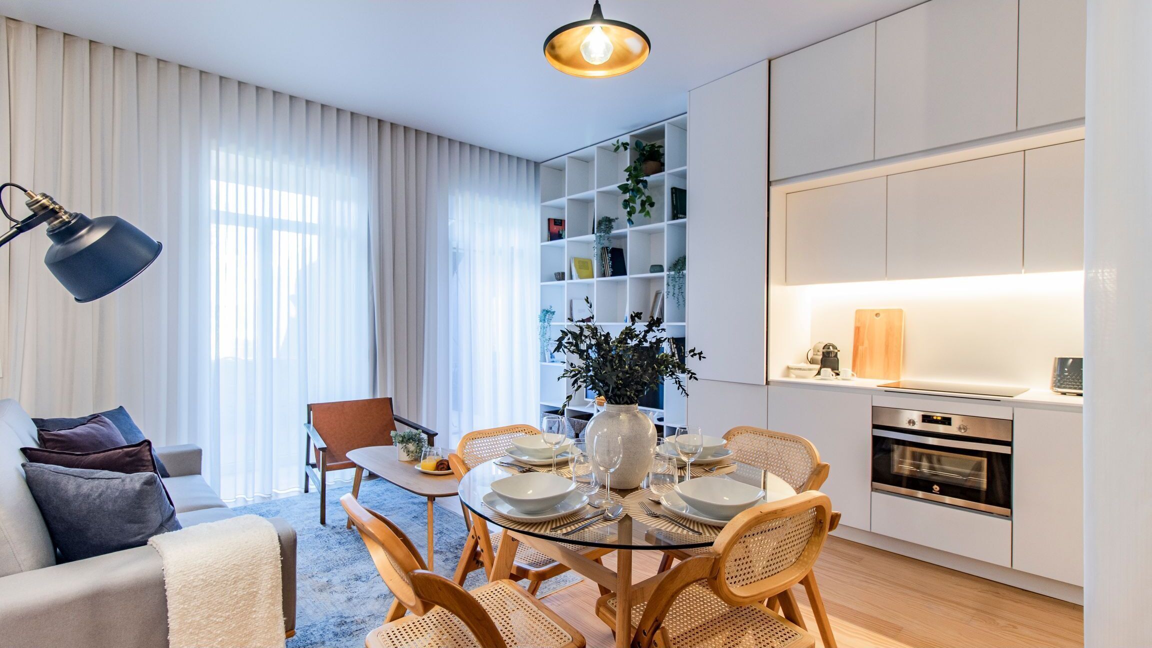 A modern living room with a glass dining table, wicker chairs, a sofa, and a bookshelf.