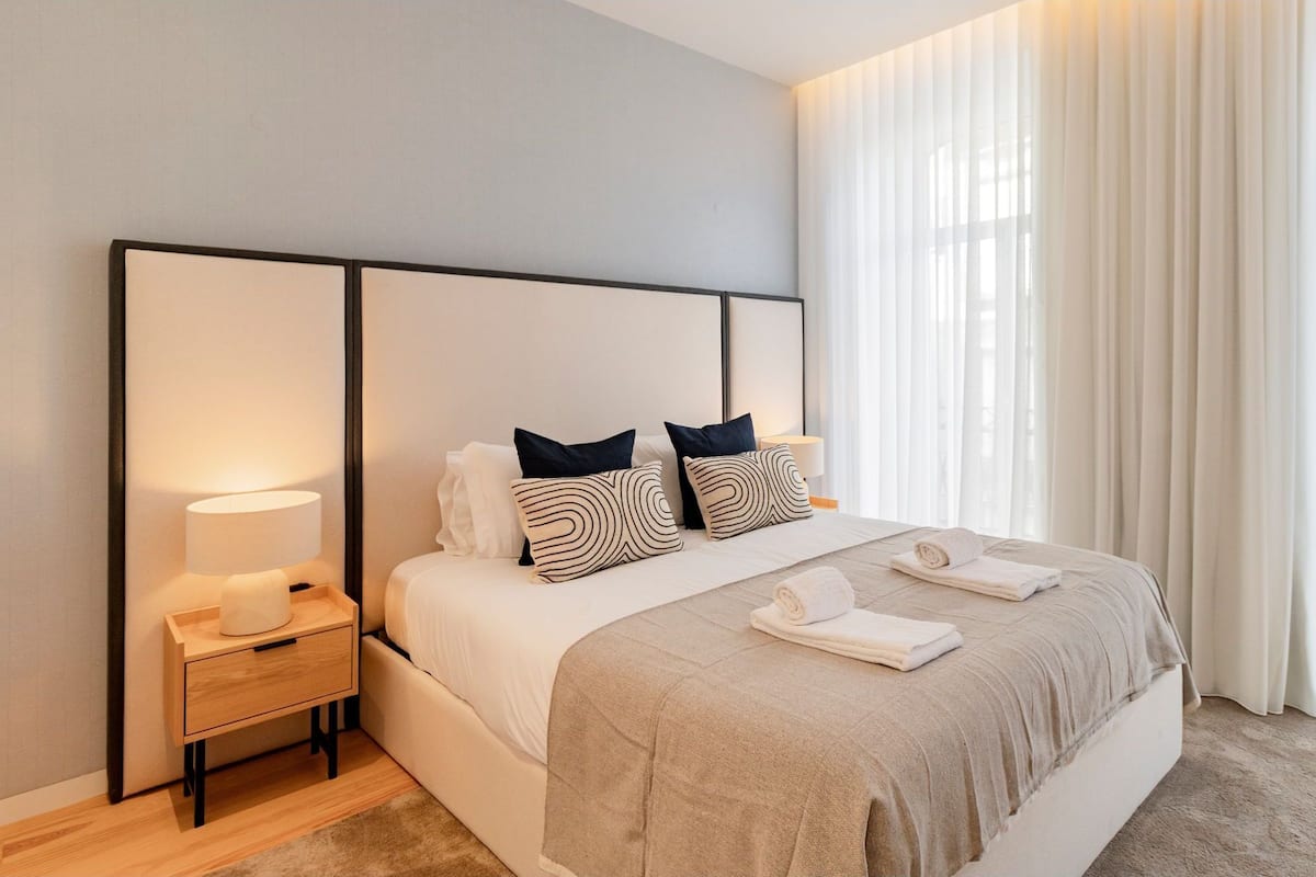Una habitación de hotel moderna con una cama grande, una mesita de noche con lámpara y cortinas traslúcidas que dejan entrar la luz natural.
