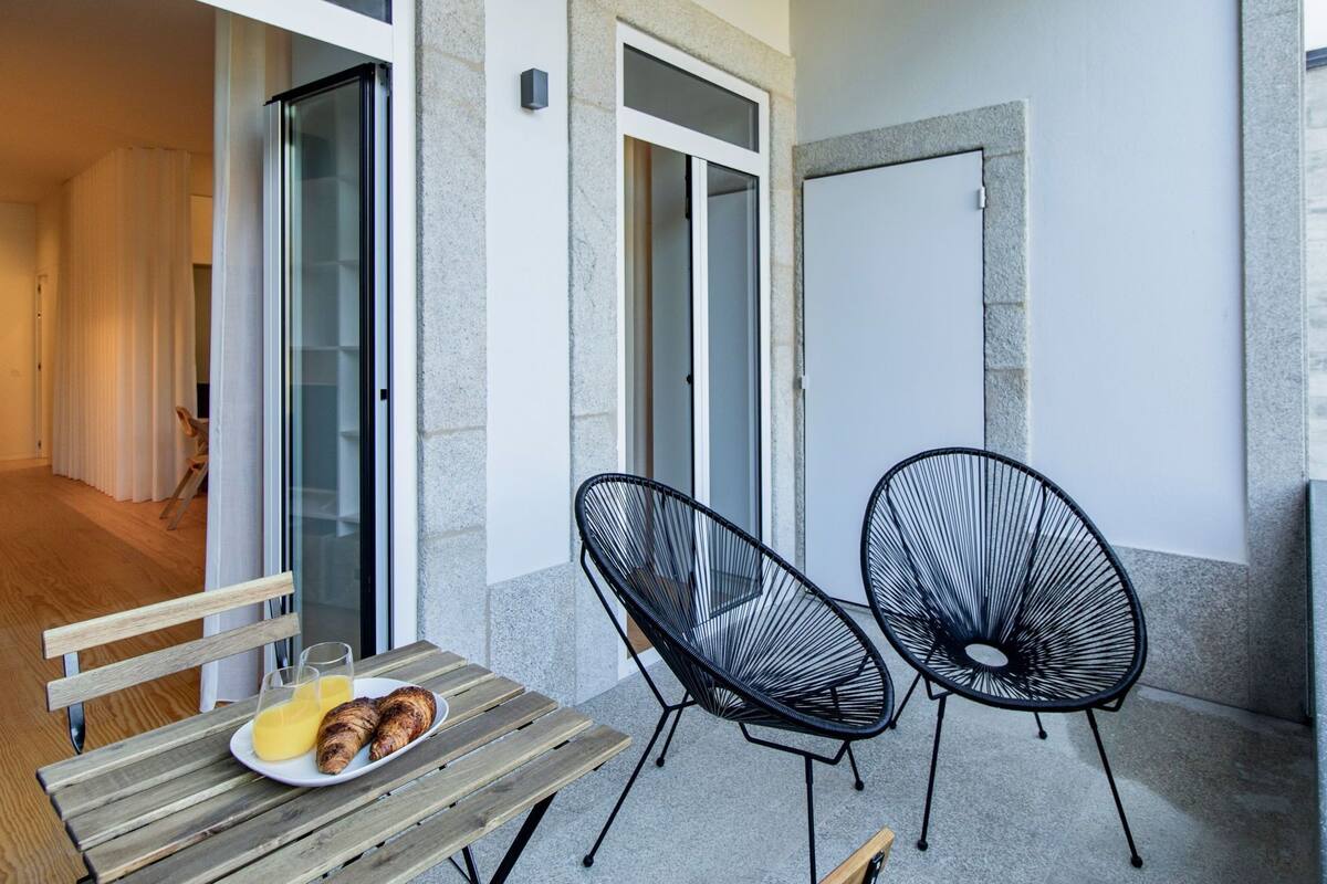 Un balcón con banca de madera, dos sillas de alambre negras y una mesa con croissants y zumo de naranja.