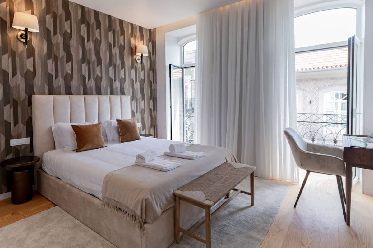 Una habitación de hotel moderna con una cama grande, un banco de madera, un escritorio y vistas de la ciudad a través de amplias ventanas.