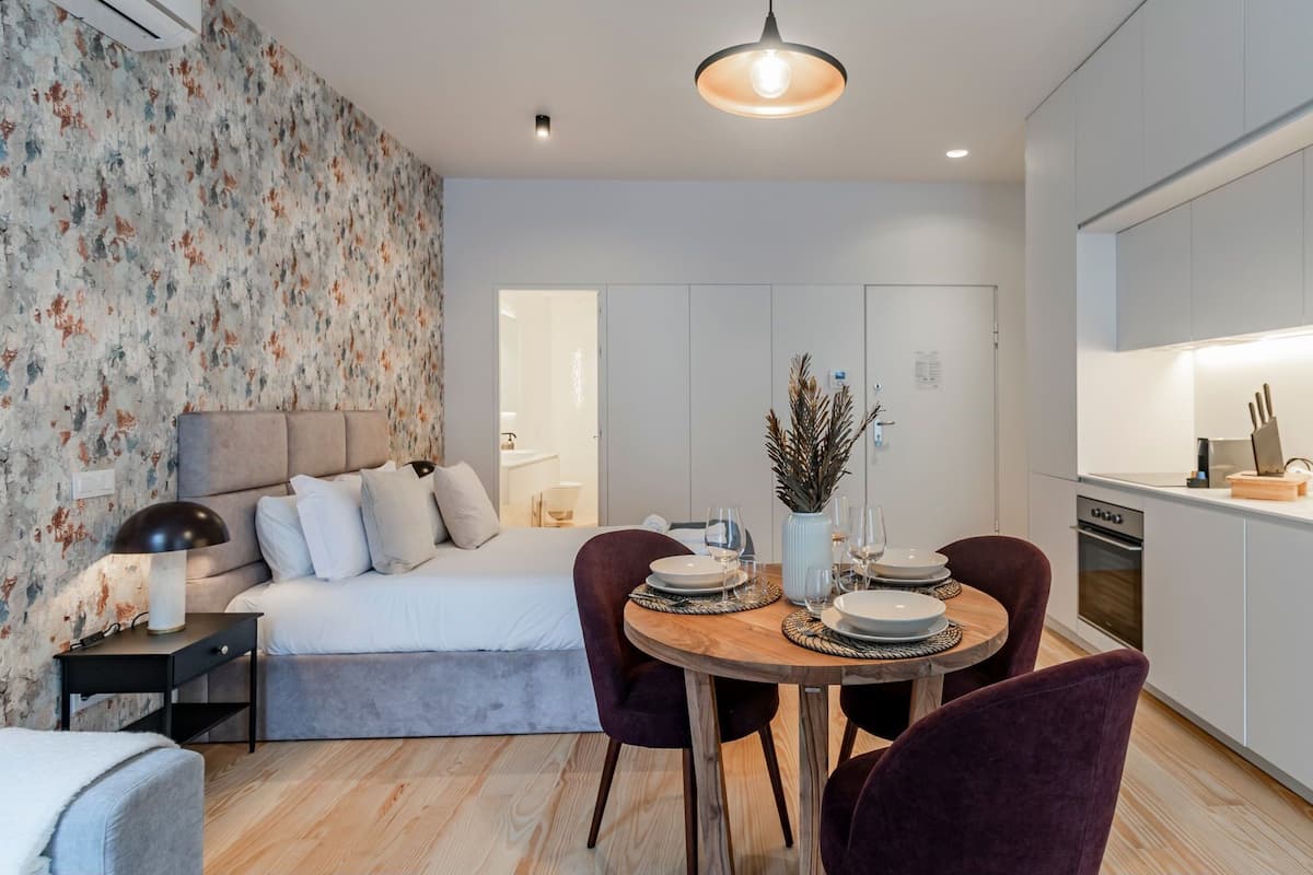 Una habitación de hotel moderna con cama, una mesa de comedor para dos personas, una zona de cocina y un baño visible a través de una puerta abierta.