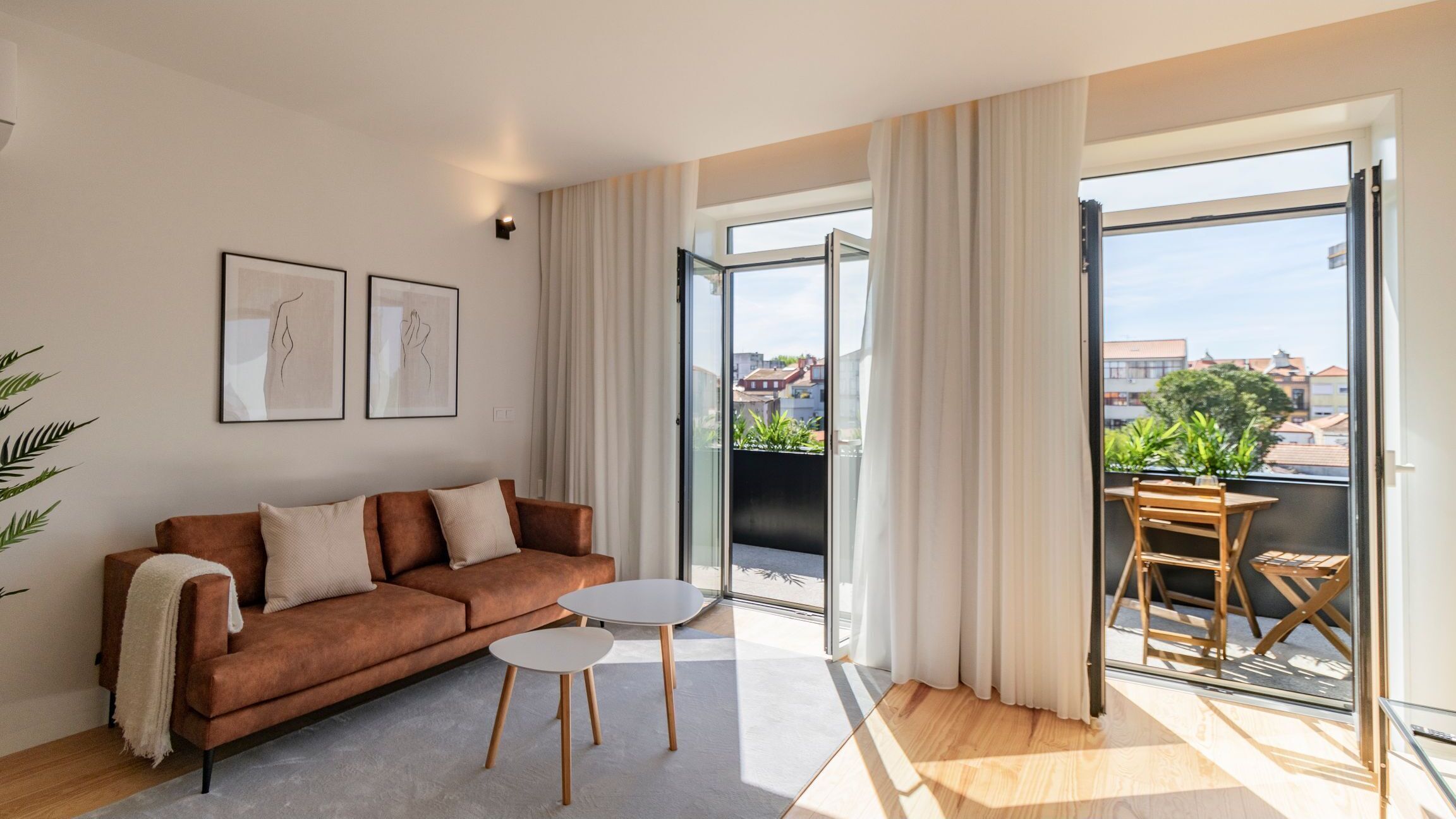 A modern living room with a brown sofa, a small round table, and a balcony with a wooden table and chairs.