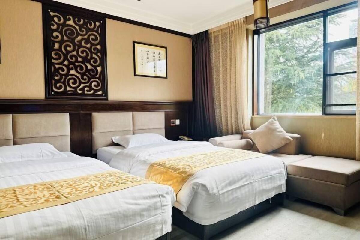 A hotel room with two beds, a sofa, a decorative wall panel, and a window with a view of trees.