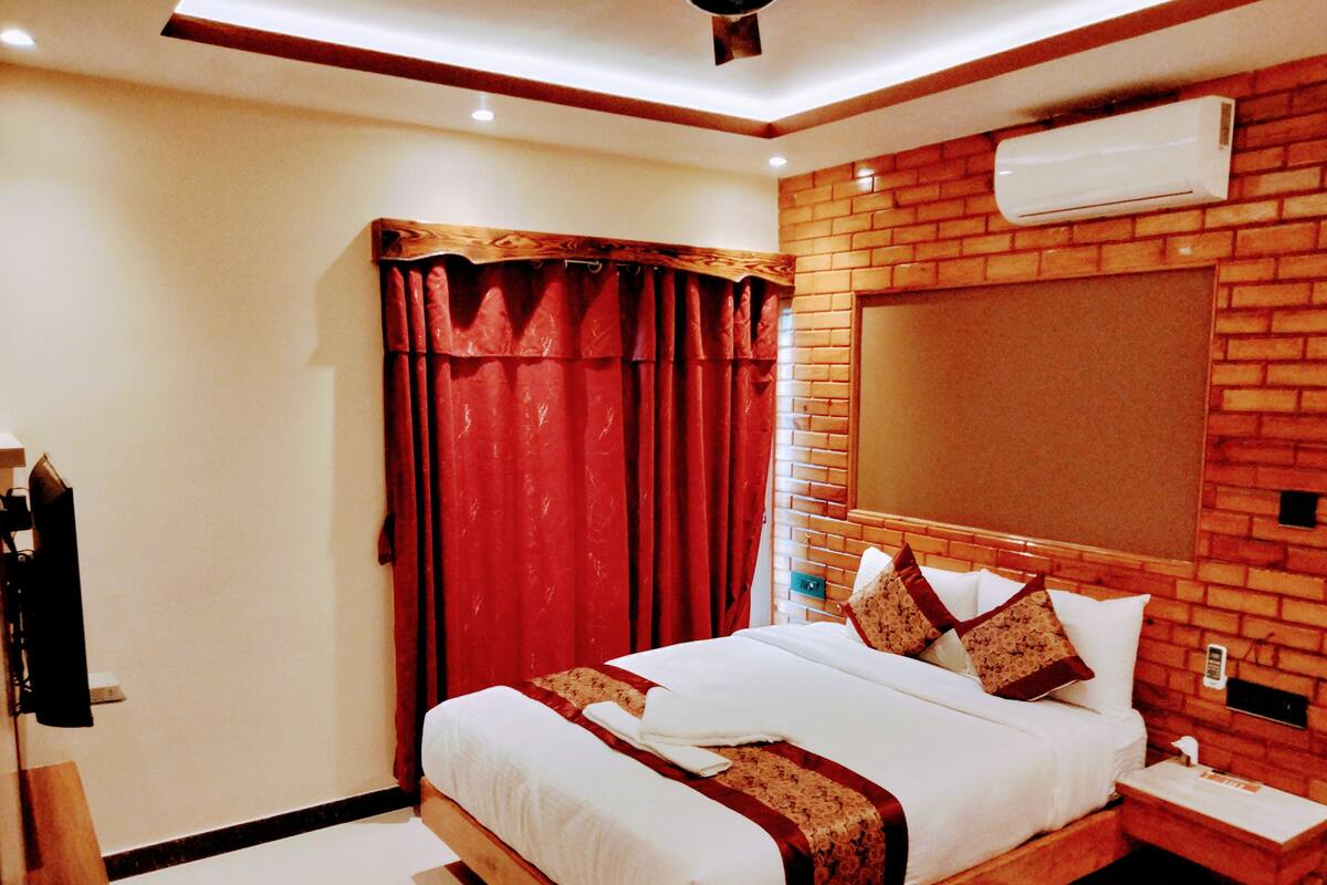A hotel room with a large bed, red curtains, a wooden headboard, and a brick-patterned wall.