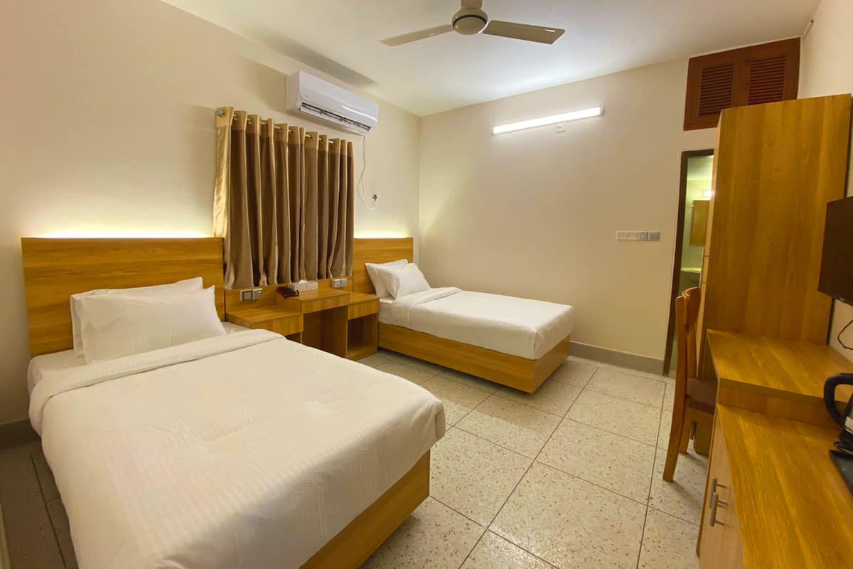 A hotel room with two beds, a ceiling fan, an air conditioning unit, a bedside table with a lamp, and a TV.