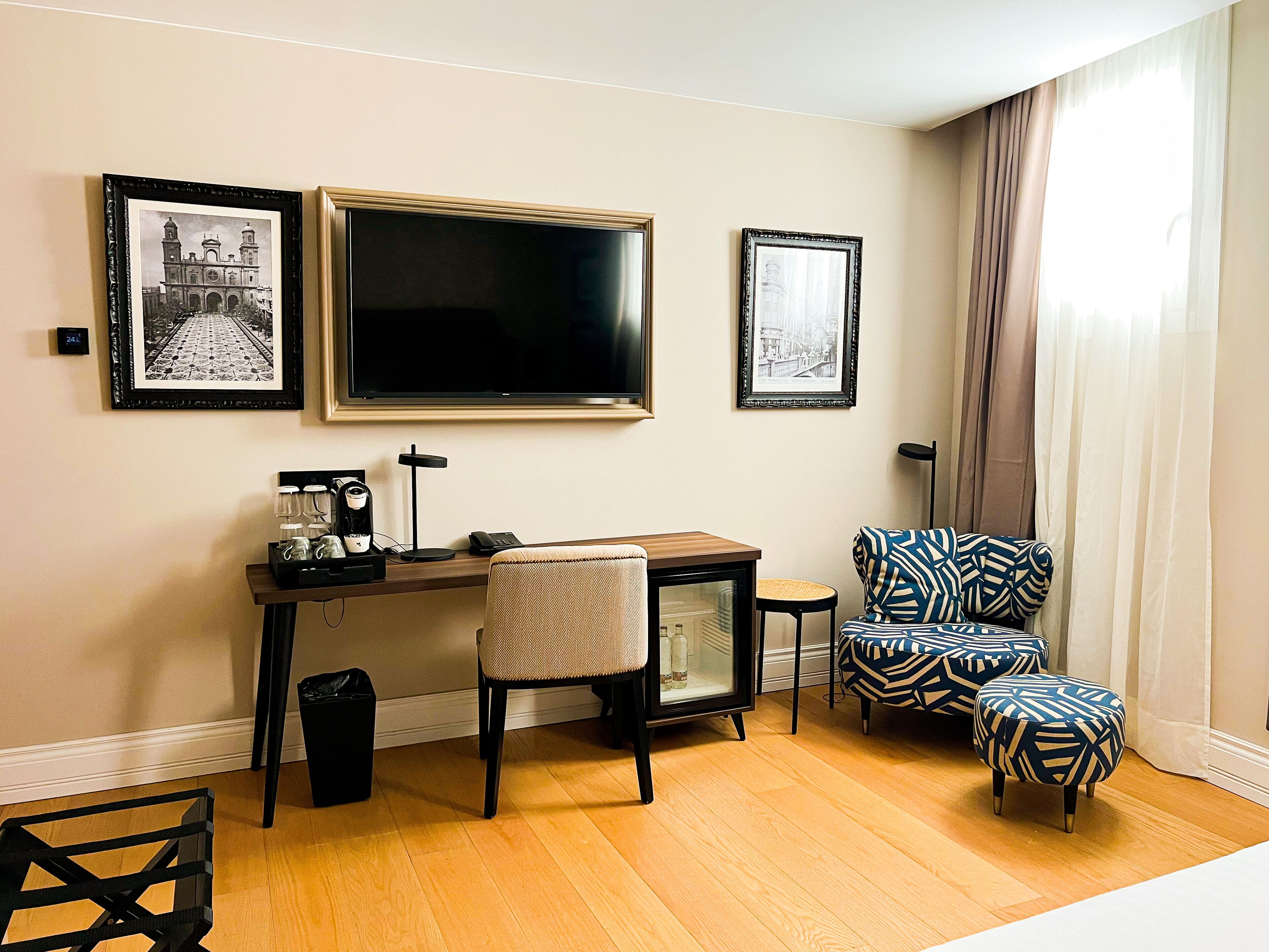 Una habitación de hotel con escritorio, silla, reposapiés y dos cuadros en la pared.