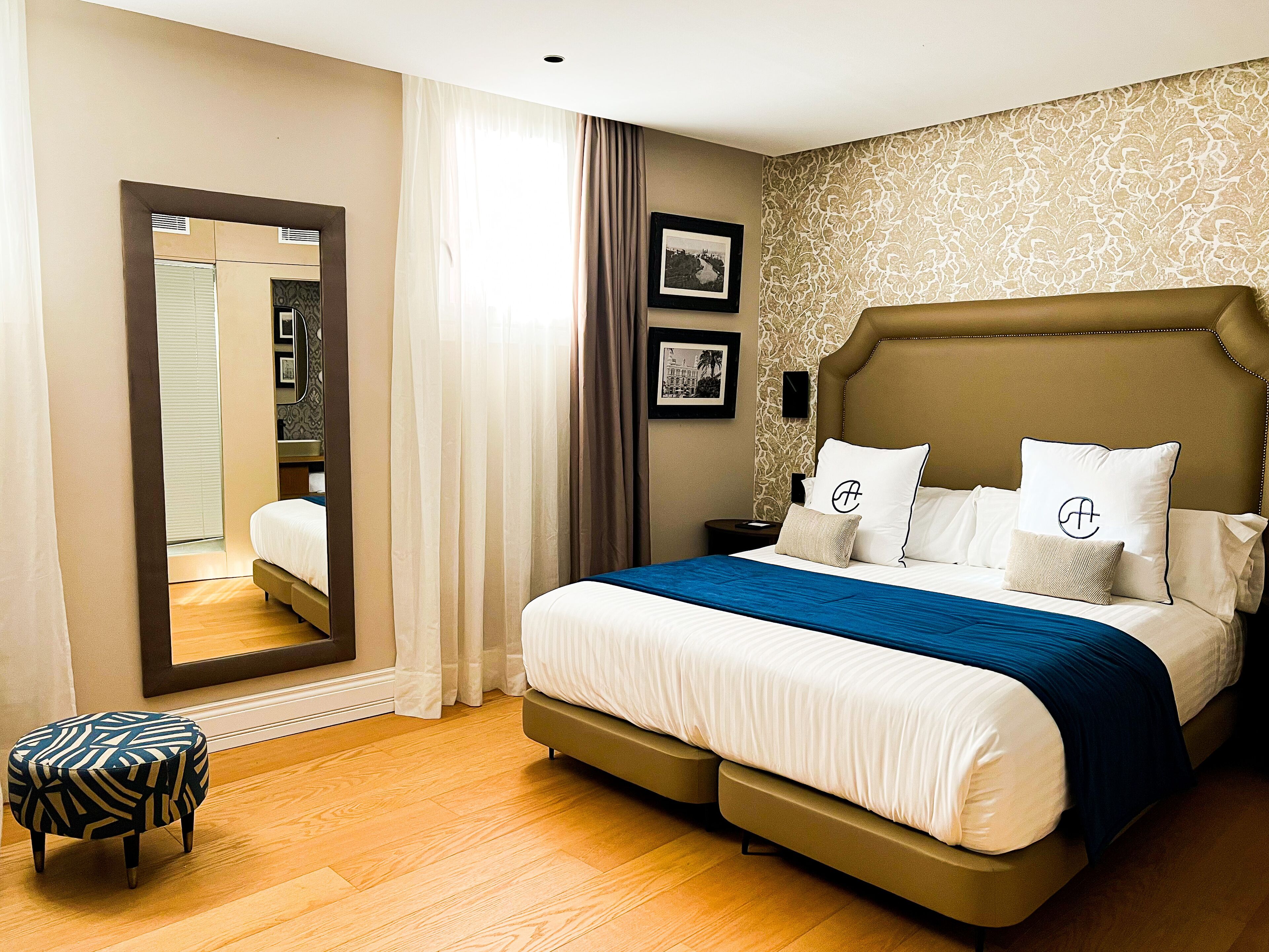 A neatly arranged hotel room with a large bed, a bedside table, a mirror, and a small stool.