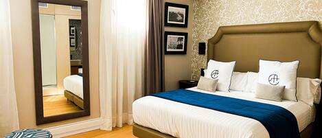 Habitación de hotel ordenada con una cama grande, una mesita de noche, un espejo y un taburete pequeño.