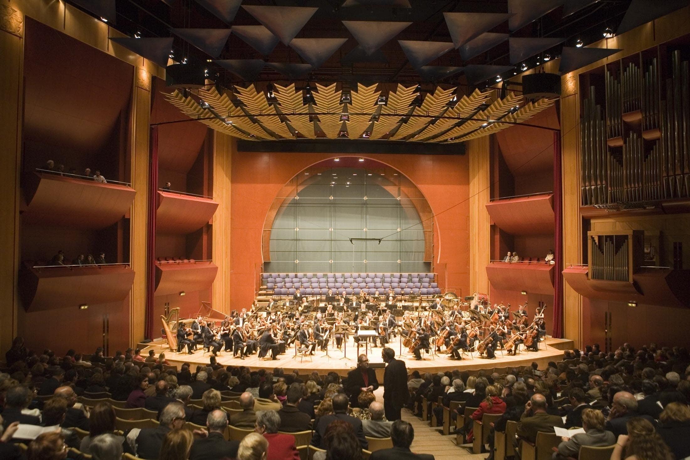 Une salle de concert avec un orchestre sur scène, un public au premier plan et un grand orgue en arrière-plan.