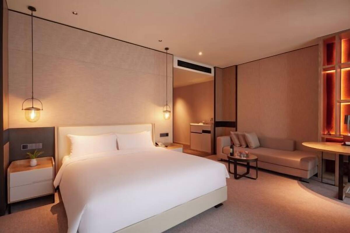 Una habitación de hotel moderna con una cama grande, mesitas de noche, un sofá, una pequeña mesa y una estantería empotrada.