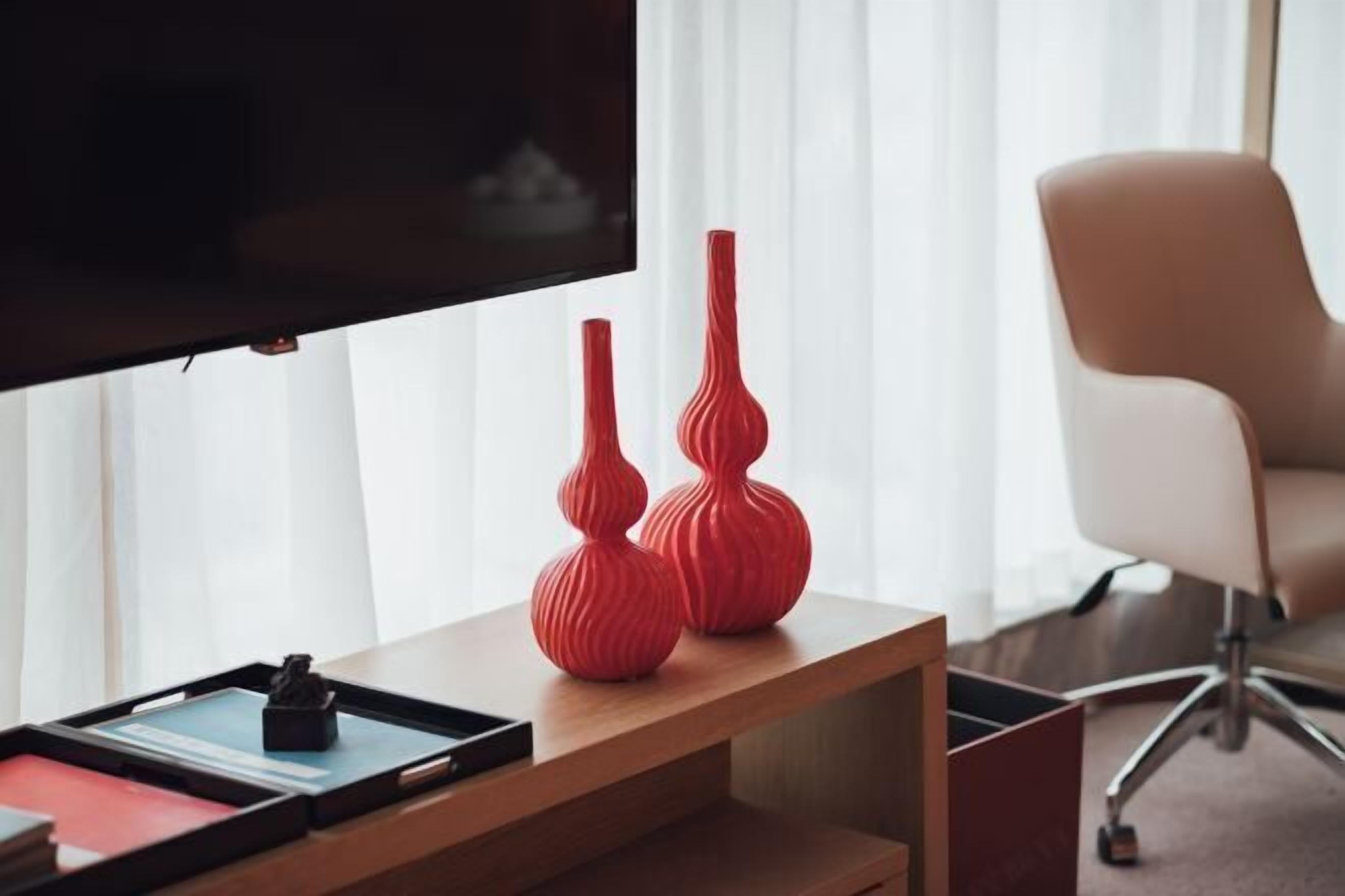 Un salon moderne avec un téléviseur à écran plat fixé au mur, deux vases rouges posés sur une table d’appoint en bois, et une chaise de bureau blanche à l’arrière-plan.