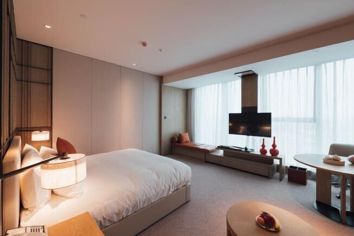 A modern hotel room with a large bed, a seating area, a dining table, and a TV.