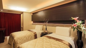 Ein Hotelzimmer mit zwei Betten, einem dunkelholznen Kopfteil, einem Nachttisch mit Blumenvase und einem roten Vorhang.