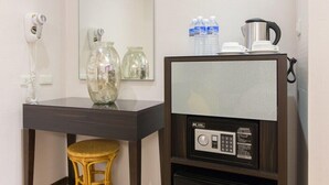 Ein Hotelzimmer mit dunklem Holztisch, einem Korbstuhl, einem Fön, einem großen Spiegel, einem Schrank mit Kaffeemaschine, Wasserflaschen und einem Glas.