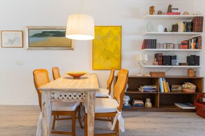 Uma área de refeições com mesa e cadeiras de madeira, um candeeiro pendente e uma estante com livros e objetos decorativos.