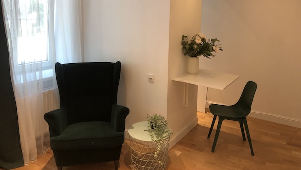 A room with a dark green armchair, a small round table with a plant, a white shelf with a vase of flowers, and a green chair.