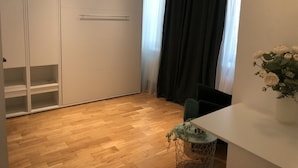 A room with a wooden floor, a white wardrobe, a small table with a vase of flowers, and a chair.