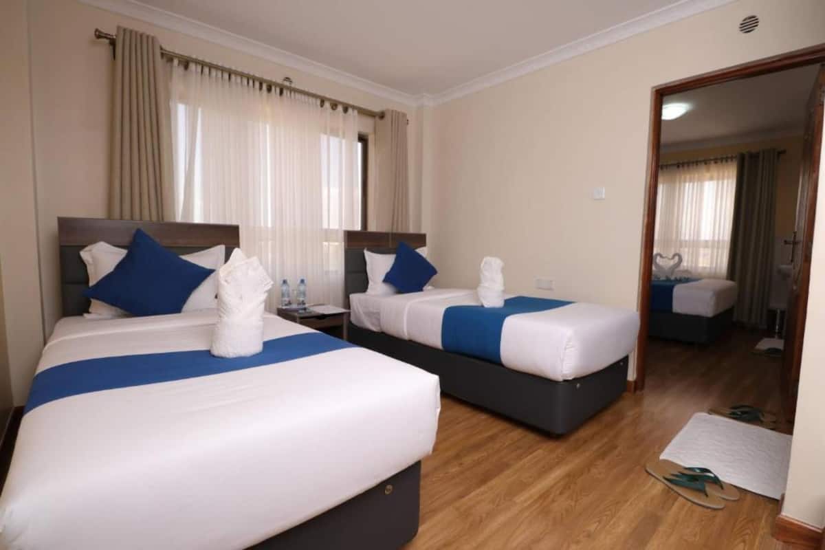 Habitación de hotel con dos camas, cada una con una almohada azul decorativa, ropa de cama blanca y una toalla sobre la cama. El suelo es de madera y hay un espejo en la pared.