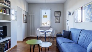 Uma sala de estar com sofá azul, uma mesa de jantar com cadeiras, uma TV em uma estante e quadros nas paredes.