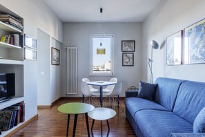 A living room with a blue sofa, a dining table with chairs, a TV on a shelf, and framed pictures on the wall.