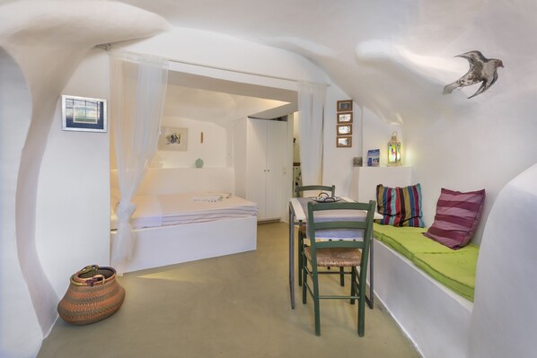 Ein kleines Zimmer mit Bett, Tisch, Stuhl und einer Bank mit Kissen.