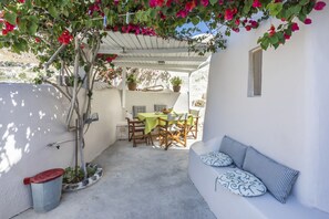 Eine überdachte Terrasse mit Tisch und Stühlen, eine Bank mit Kissen sowie eine hängende Pflanze mit roten Blüten.