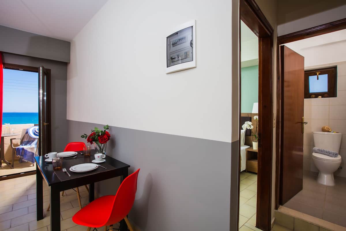 Un coin repas avec une table dressée pour deux personnes, une chaise rouge et une vue sur la mer par une porte ouverte.