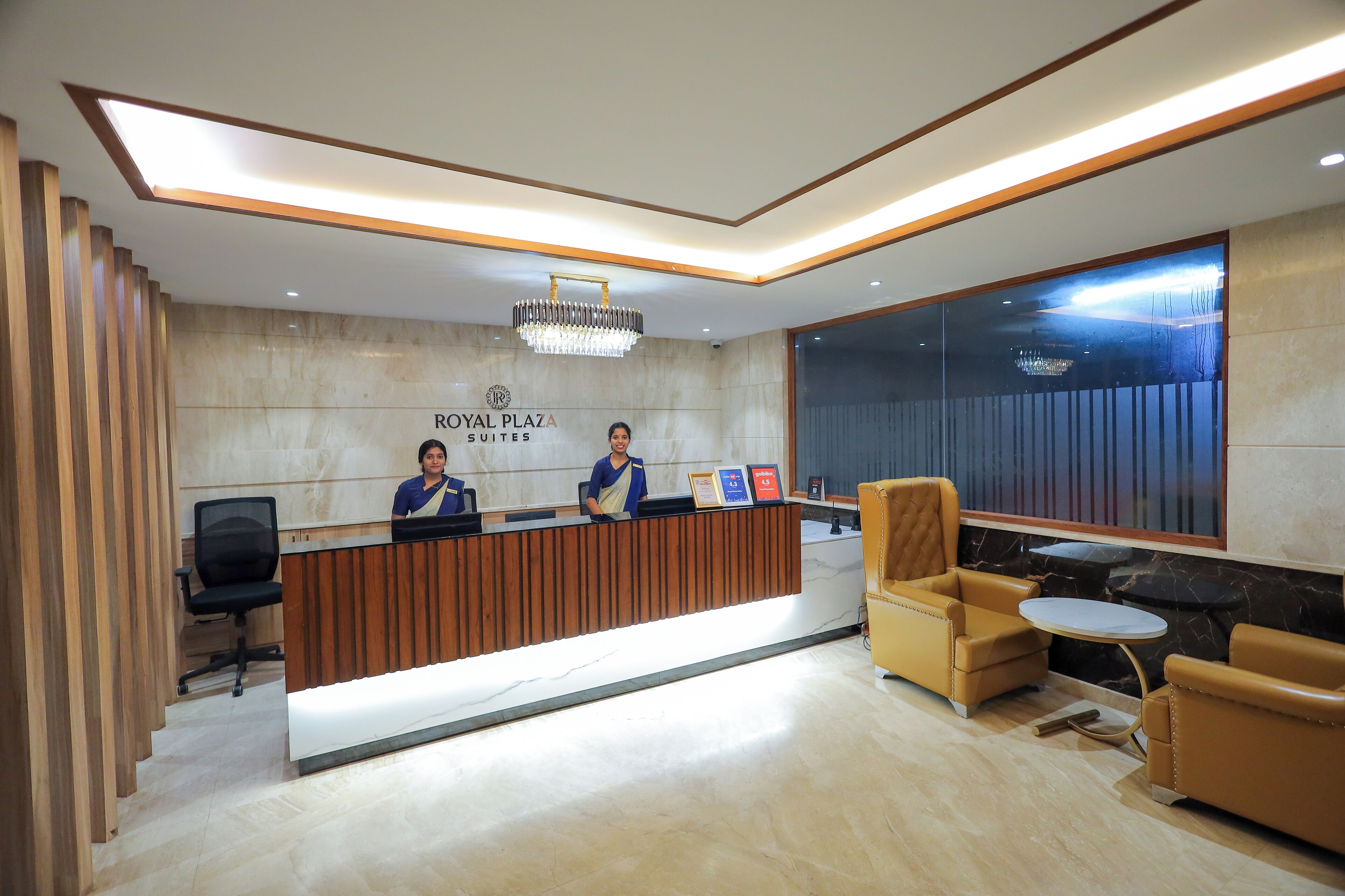 A hotel reception area with a reception desk, two receptionists, a chandelier, a seating area with a sofa and a small table, and a large window.